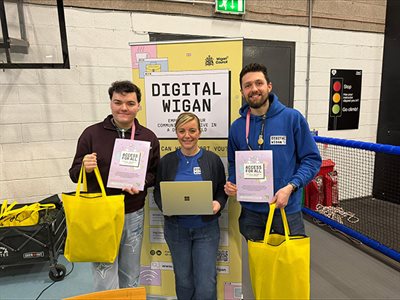 Three people stand by a Digital Wigan banner, holding “Access for All” leaflets and yellow bags.