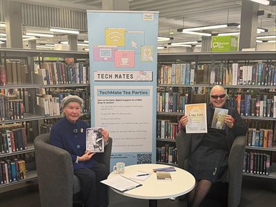 Two people sit in a library beside a Tech Mates banner, holding books at a small table with materials for a digital support session.