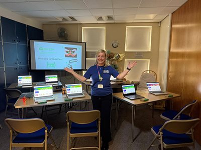 Presenter stands in classroom with laptops, displaying a “Welcome to Code Club” micro:bit lesson on a large screen.