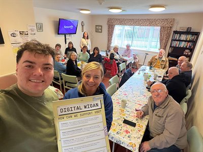 Group seated around tables in a community room, two people holding “Digital Wigan” sign, others using laptops and chatting.