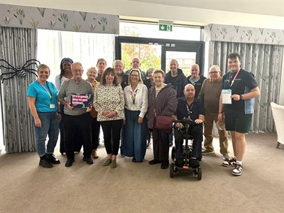 Group gathered in community room, one person holding a sign reading &amp;quot;It&amp;#39;s a good day to get online!&amp;quot;, another using a mobility scooter, staff standing alongside.