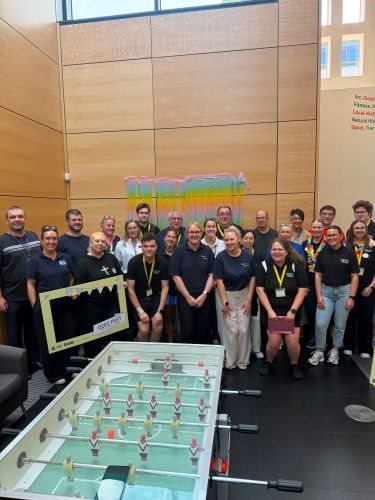Group of people all stood behind a table football game looking at the camera. They are all wearing Lanyards that say digi fest on them and some people are holding photo booth props