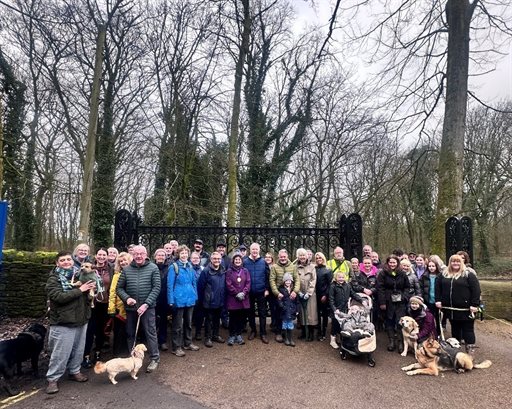 A large group of people stand together for a group photo in an outdoor park setting, positioned in front of an ornate black gate surrounded by tall, leafless trees. Many individuals are wearing coats and outdoor clothing suited for cold weather. Several d