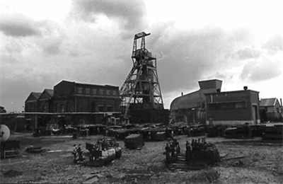 A black‑and‑white photograph of an industrial mining site. A tall steel headframe tower stands at the centre, surrounded by several large brick and metal buildings. The ground is cluttered with equipment, machinery parts, and scattered materials. The sky