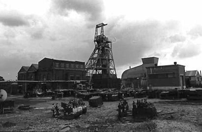 A black‑and‑white photograph of an industrial mining site. A tall steel headframe tower stands at the centre, surrounded by several large brick and metal buildings. The ground is cluttered with equipment, machinery parts, and scattered materials. The sky