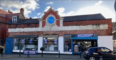 A wide, single‑storey brick building with a decorative gabled centre section featuring a circular blue YMCA sign high on the façade. The ground floor has been modernised and houses a “Bathroom Clearance Outlet,” with large display windows showing bathroom