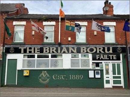 A contemporary picture of the Brian Borou Irish Club. A two storey red brick building, decorated with flags. Four windows on the second storey, a large sign showing the name ‘Brian Borou’ across the fascia and a paining of an Irish Harp with the words. Es