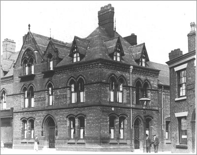 Black and white photo of Leigh Lilford hotel. The building has arched windows and doorways and decorative stonework. A traditional streetlamp stands by the corner, and a few pedestrians in period clothing are visible on the pavement.