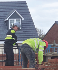 Two people wearing hi-vis jackets and hard hats on a construction site near a brick wall