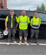 Three men in hi-vis jackets, one of them holding a hard hat all stood by a black van