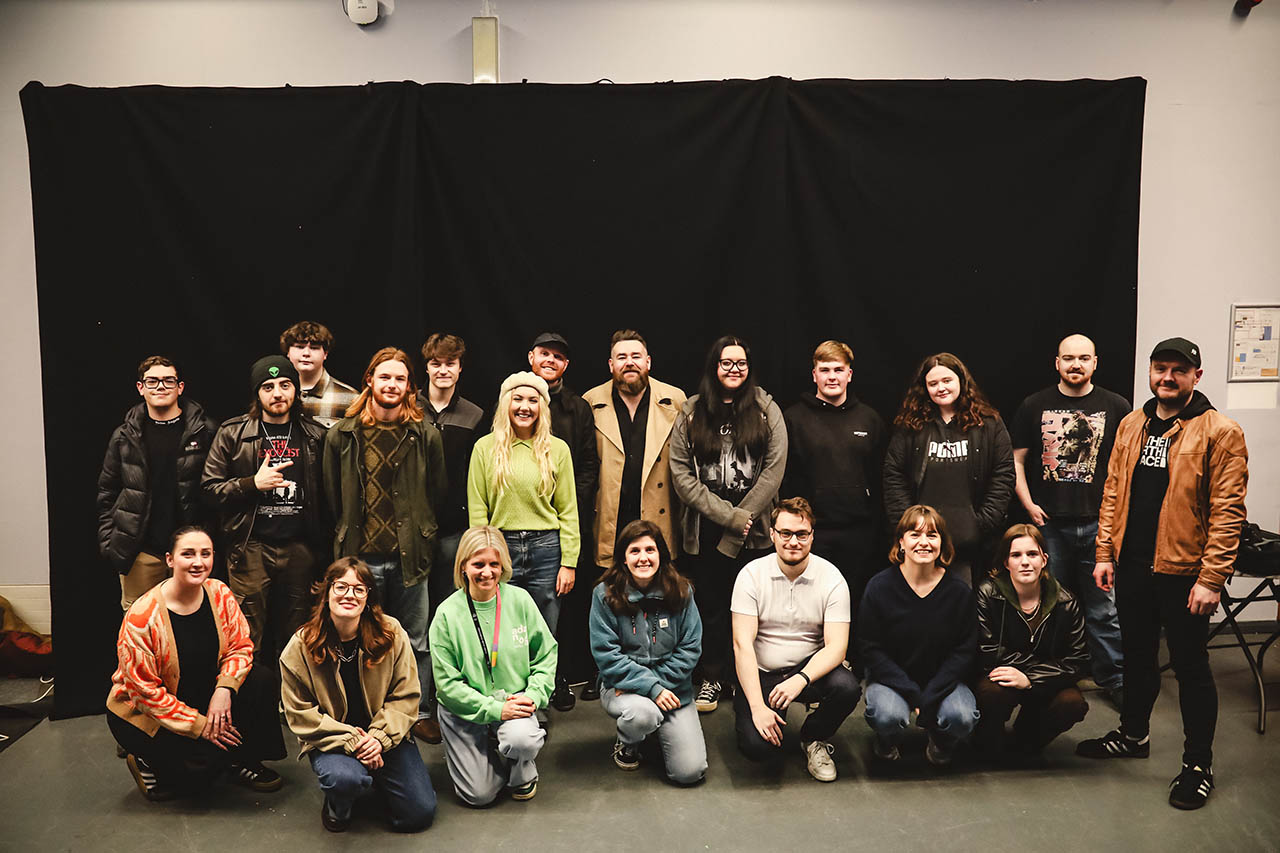 People in casual clothes pose in two rows indoors against a black backdrop, with some kneeling in front.