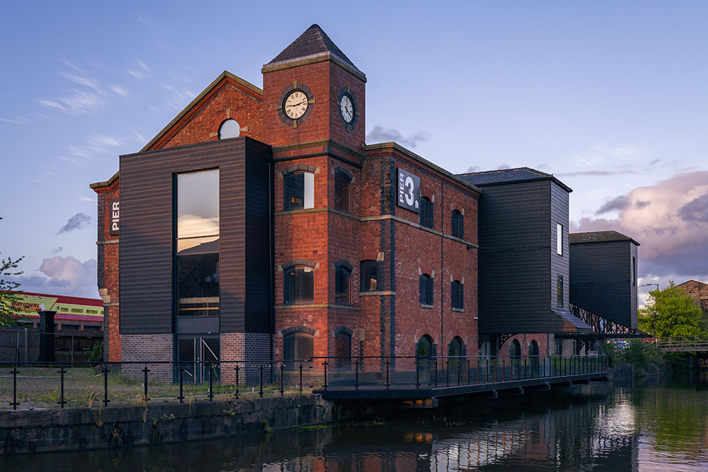 Historic canal-side brick building with clock tower and modern extensions reflected in calm water at dusk.