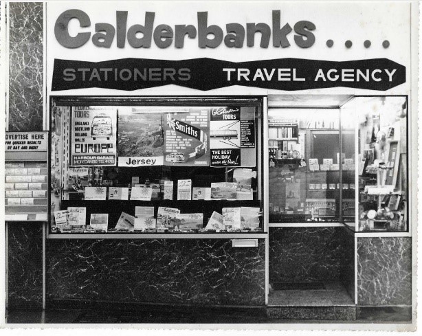 Black‑and‑white photograph of the Calderbanks shopfront, featuring signage for &lsquo;Stationers&rsquo; and &lsquo;Travel Agency.&rsquo; The display window is filled with travel posters advertising destinations such as Europe and Jersey, along with brochures and pamphlets arrang