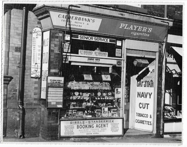 Black and white photograph of the Calderbank&rsquo;s shopfront, displaying a small tobacconist and booking office. The windows are filled with cigarette packets, postcards and small goods. Signs for &lsquo;Player&rsquo;s Cigarettes,&rsquo; &lsquo;Senior Service,&rsquo; and &lsquo;Navy Cut Tobacco