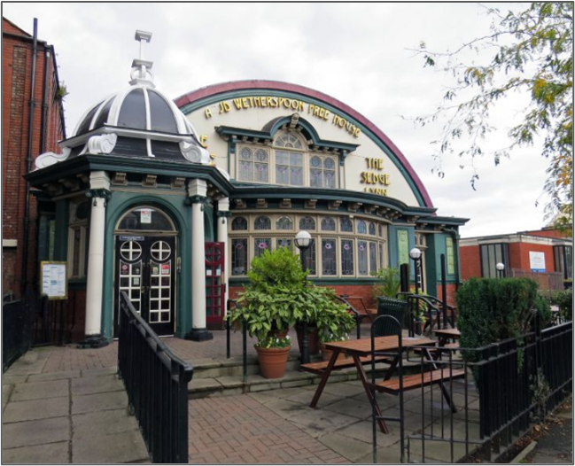 A historic building with an arched roofline, ornate windows, and a domed entrance porch, featuring green and cream detailing. The foreground shows outdoor seating with wooden benches, potted plants, and a paved path leading to the main door.