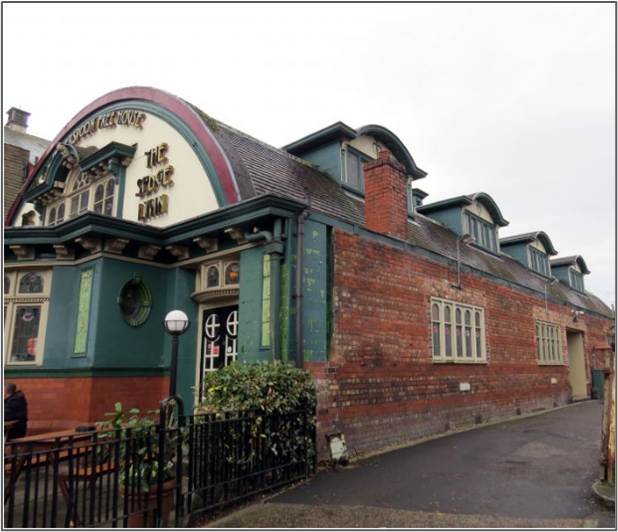 A side view of a brick building with green and cream decorative detailing, arched attic windows, and a rounded gabled entrance section bearing the sign &ldquo;The Sedge Lynn.&rdquo; A narrow pavement runs alongside the building, with shrubs and a fenced area near the