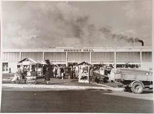 Black and while image of Leigh Markets in the 1970s. Wooden stalls outside the market hall, smoke coming out of the chimney and lots of people walking around the outdoor markets