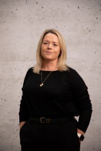 Person with blonde, shoulder‑length hair standing against a plain background, wearing a black long-sleeve top with hands in pockets