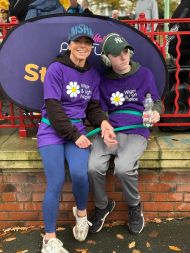 Joanne and her son Harry sat close together on a low brick wall at an outdoor event. Both are wearing matching purple T‑shirts with a large white and yellow flower graphic and the words &ldquo;Wish Upon a Star&rdquo; printed on the front. Joanne is wearing blue leggi