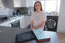 Leanne sat on a stool in a modern kitchen, wearing a white T‑shirt with a circular logo that reads &ldquo;Clever Cookie School of Tutoring.&rdquo; In front of them on the countertop are a black drawstring bag with the same logo and a closed box with a teal and red de