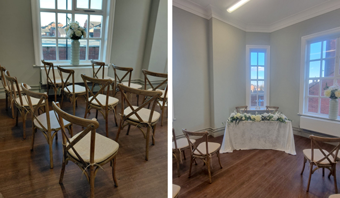 Wedding ceremony setup with a white-draped table decorated with floral arrangements, placed in front of tall windows. Wooden chairs with cream cushions are arranged around the table.