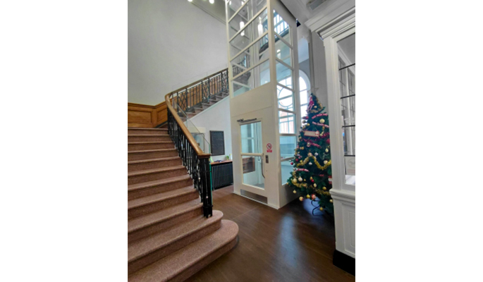 Building foyer with a wooden staircase, next to a modern glass lift. A decorated Christmas tree with gold and red ornaments stands beside the lift. Large arched windows allow natural light into the space.